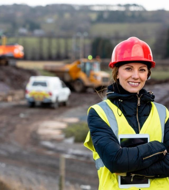 A portrait of a woman, part of a survey crew looking at the camera and smiling with her arms crossed while wearing a hardhat and reflective clothing. She is working in the North East of England on a green field site and holding a digital tablet. The area where she is working is behind her.